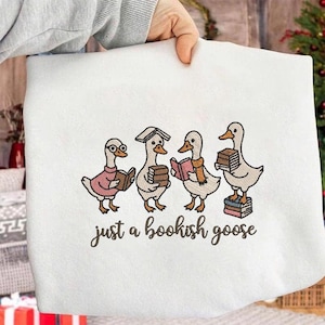 May include: White sweatshirt with an embroidered design of four cartoon geese, each holding books. The geese wear glasses, sweaters, and scarves. The text "just a bookish goose" is embroidered below the geese.