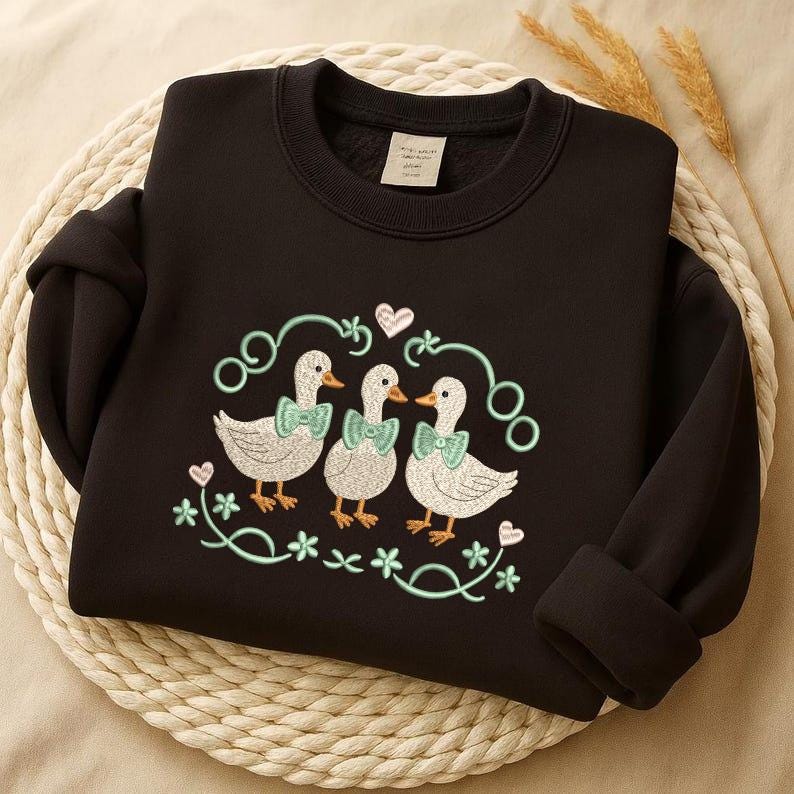 May include: Black sweatshirt with an embroidered design of three ducks wearing bow ties. The ducks are surrounded by a floral and heart-shaped design in light green. The sweatshirt is folded on a woven mat.