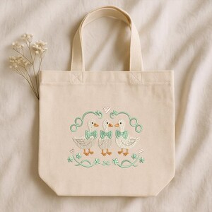 May include: Cream canvas tote bag with an embroidered design of three ducks wearing bow ties. The ducks are surrounded by green swirls, hearts, and floral accents. The bag has a rectangular shape with a handle.