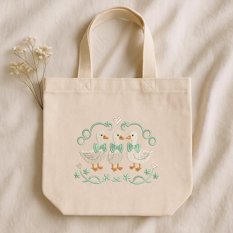 May include: Cream canvas tote bag with an embroidered design of three ducks wearing bow ties. The ducks are surrounded by green swirls, hearts, and floral accents. The bag has a rectangular shape with a handle.
