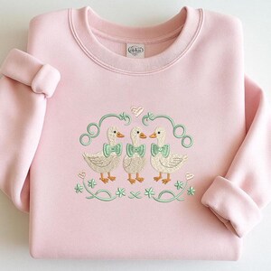 May include: A pale pink sweatshirt with a crew neck and turned-up sleeves. The front displays an embroidered design of three ducks wearing green bow ties, framed by green swirls, hearts, and shamrocks. The brand name "Gildan" is visible on the inside collar.