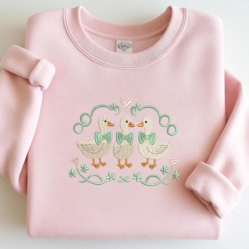 May include: A pale pink sweatshirt with a crew neck and turned-up sleeves. The front displays an embroidered design of three ducks wearing green bow ties, framed by green swirls, hearts, and shamrocks. The brand name "Gildan" is visible on the inside collar.
