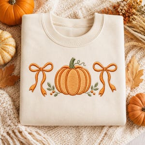 May include: A cream-colored crewneck sweatshirt featuring an embroidered design of a central orange pumpkin flanked by two orange ribbons and small green leafy sprigs with tiny orange berries on each side of the pumpkin.