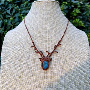 May include: A brown macrame necklace featuring a deer antler design. The pendant holds a blue-green oval gemstone. The necklace is displayed on a mannequin with a neutral background.