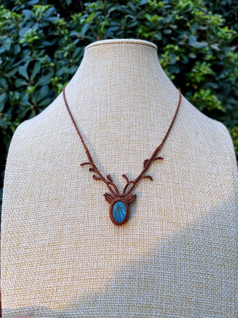 May include: A brown macrame necklace featuring a deer antler design. The pendant holds a blue-green oval gemstone. The necklace is displayed on a mannequin with a neutral background.