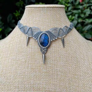 Peut inclure: Un collier ras du cou en macramé gris avec une pierre précieuse ovale bleue au centre et des pendentifs en forme de pic argentés. Le collier est présenté sur un mannequin, mettant en valeur son design tissé complexe et ses couleurs contrastées.