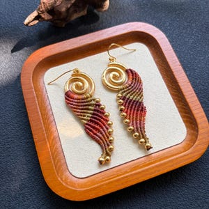 May include: A pair of dangle earrings featuring a gold-tone spiral top connected to a macrame wing-shaped drop. The macrame section is crafted from variegated thread in shades of red, orange, and olive green, accented with small gold-tone beads.