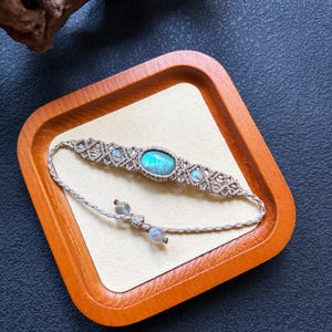 Op de afbeelding: Een macramé armband met een centrale ovale blauwe maansteen, twee kleinere ronde maanstenen en ingewikkeld beige koordvlechtwerk. De verstelbare uiteinden van het koord zijn afgewerkt met twee kleine ronde kralen voor een gedetailleerd ontwerp.
