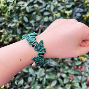 May include: A green macrame bracelet with a leaf design and black beads. The bracelet is made of knotted cord and is worn on a wrist. The background is blurred green foliage.