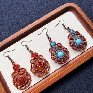 May include: Four pairs of handmade macrame earrings displayed on a wooden tray. The earrings feature intricate woven patterns in shades of brown, with accents of red and turquoise gemstones. The earrings have a boho style and are perfect for adding a unique touch to any outfit.