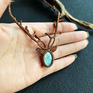 May include: A brown macrame necklace featuring a deer antler design. The pendant holds a blue-green oval stone. The necklace is displayed on a hand against a dark background, showcasing the intricate knotting.