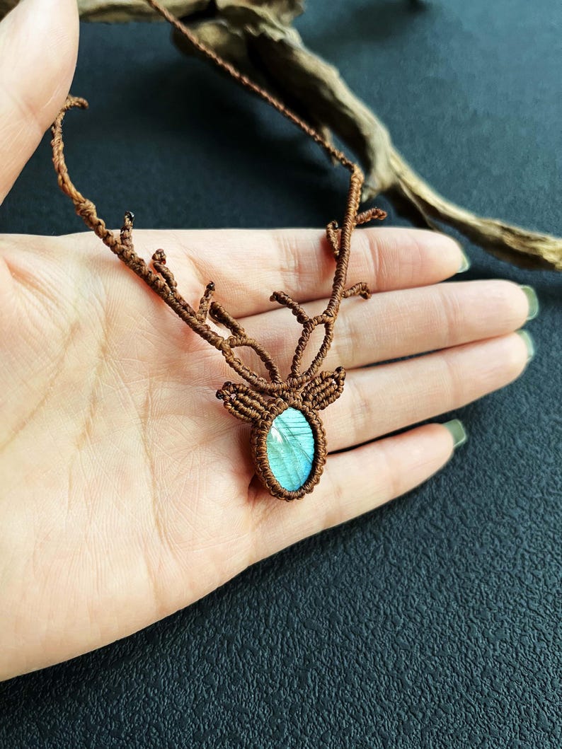 May include: A brown macrame necklace featuring a deer antler design. The pendant holds a blue-green oval stone. The necklace is displayed on a hand against a dark background, showcasing the intricate knotting.