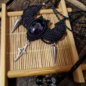 Könnte beinhalten: Handgefertigte schwarze Makramee-Halskette mit einem großen, ovalen, lila Edelstein in der Mitte. Die Halskette hat silberfarbene Kegel- und sternförmige Anhänger. Das aufwendige Design wird vor einem Bambushintergrund präsentiert, der den einzigartigen Stil des Schmucks hervorhebt.