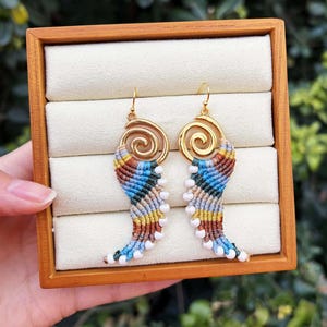Puede incluir: Un par de pendientes hechos a mano con una parte superior en espiral dorada y un diseño de macramé colorido. El macramé presenta hilos azules, amarillos, marrones y blancos. Los pendientes se muestran en una caja de joyería de madera.