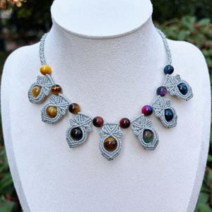 Puede incluir: Un collar hecho a mano con un cordón de macramé gris claro y cuentas de piedras preciosas de colores. El collar tiene elementos decorativos de macramé, cada uno con una piedra preciosa de diferente color, incluyendo amarillo, marrón y azul. Se exhibe en un maniquí blanco.