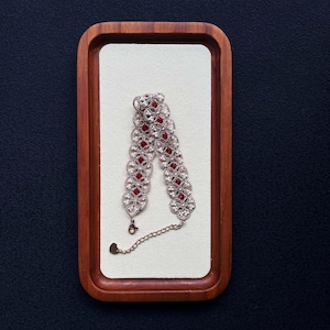 May include: A silver bracelet with red gemstones and a heart-shaped charm. The bracelet is displayed on a white surface inside a wooden tray, set against a dark background. The bracelet features a delicate, ornate design.