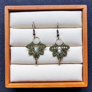 May include: A pair of handmade macrame earrings displayed in a wooden box. The earrings feature a leaf-like design in olive green with a small, round, light blue stone in the center. The earrings have gold-colored accents and a small gold teardrop charm.