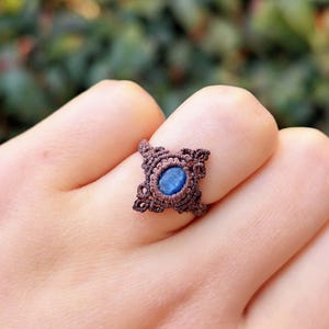 Può includere: Anello in macramè marrone fatto a mano con un design quadrato e una gemma blu ovale al centro. L'anello è realizzato con cordoncino marrone, mostrando nodi intricati. Un gioiello unico.