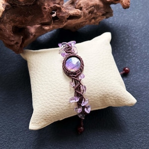 May include: A handcrafted macrame bracelet featuring a central, round, iridescent stone in shades of purple and blue. The bracelet is adorned with small, clear, purple beads and a dark brown macrame cord. The bracelet rests on a cream-colored pillow.