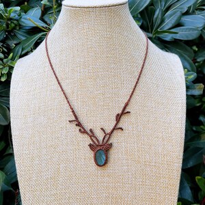 May include: A brown macrame necklace with a pendant featuring a blue-green oval stone. The necklace has a deer antler design. The necklace is displayed on a beige mannequin against a backdrop of green foliage.
