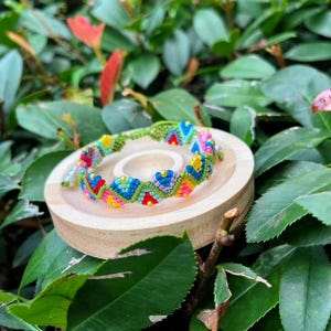 Könnte beinhalten: Ein farbenfrohes Makramee-Armband mit einem sich wiederholenden geometrischen Muster in Blau, Gelb, Rosa und Rot. Das Armband wird auf einem kleinen, runden Holzständer präsentiert, umgeben von grünem Laub. Das Design des Armbands ist aufwendig und auffällig.