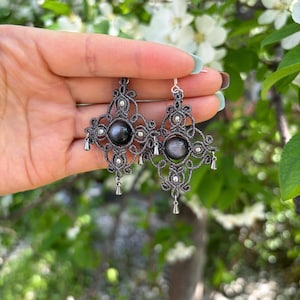 Puede incluir: Un par de pendientes de macramé gris oscuro con una cuenta central gris oscuro y pequeños detalles plateados. Los pendientes tienen un diseño geométrico con colgantes de plata en forma de lágrima. Se muestran sobre un fondo de hojas verdes y flores blancas.