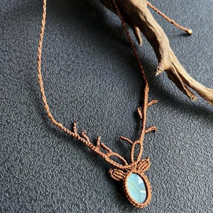 May include: A brown macrame necklace featuring a deer antler design. The pendant holds a blue-green oval gemstone. The necklace is displayed on a dark gray textured surface, with a piece of driftwood in the background.