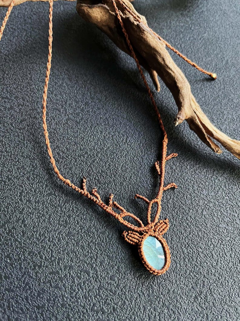 May include: A brown macrame necklace featuring a deer antler design. The pendant holds a blue-green oval gemstone. The necklace is displayed on a dark gray textured surface, with a piece of driftwood in the background.