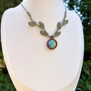 Puede incluir: Un collar de macram&eacute; con un colgante de piedra labradorita redonda con iridiscencia azul y amarilla, engastado en cord&oacute;n marr&oacute;n. La banda del collar est&aacute; hecha de cord&oacute;n verde oliva y decorada con detalles de macram&eacute; en forma de hoja.