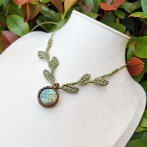 Puede incluir: Un collar de macram&eacute; con una piedra de labradorita redonda con un borde de cord&oacute;n marr&oacute;n oscuro. El cord&oacute;n del collar es de color verde oliva claro e incorpora una serie de peque&ntilde;os detalles tejidos en forma de hoja que salen del engaste central.