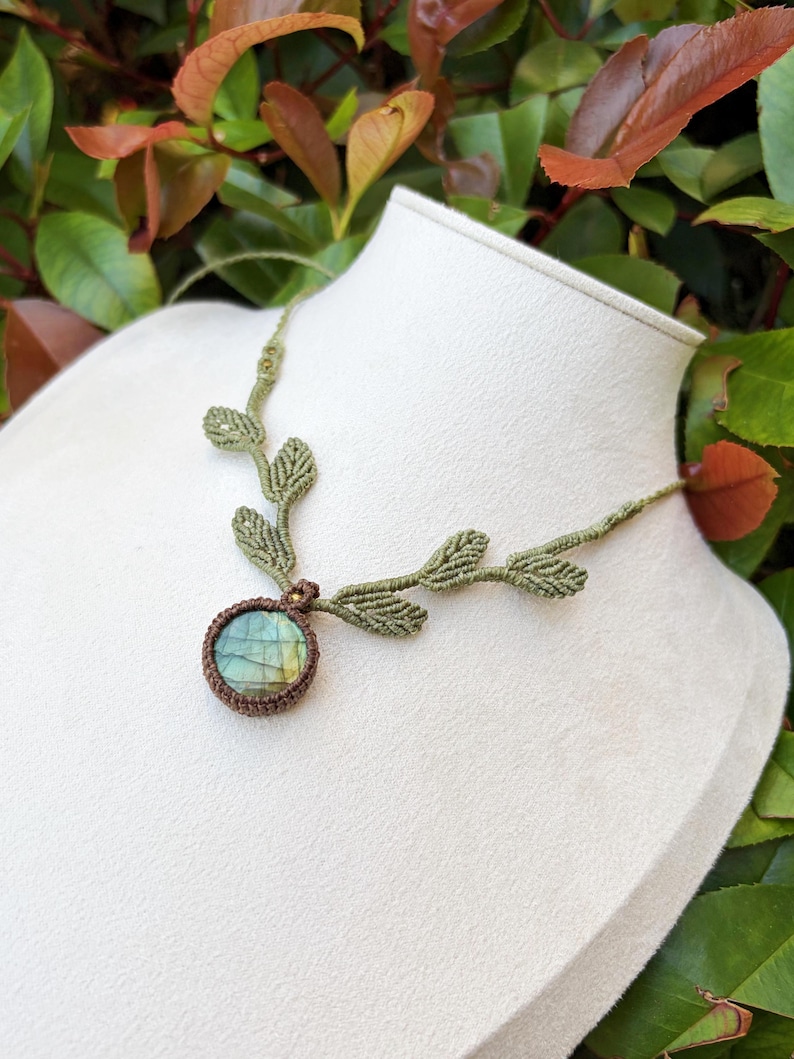 Puede incluir: Un collar de macram&eacute; con una piedra de labradorita redonda con un borde de cord&oacute;n marr&oacute;n oscuro. El cord&oacute;n del collar es de color verde oliva claro e incorpora una serie de peque&ntilde;os detalles tejidos en forma de hoja que salen del engaste central.