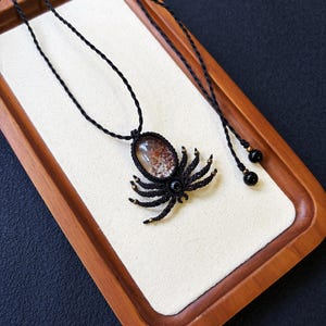 May include: A spider-shaped pendant necklace crafted from black cord using macrame techniques. The spider's body features an oval-shaped translucent stone with reddish-brown inclusions, accented by small metallic beads on the legs and black beads.