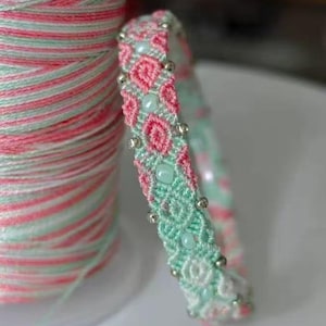 Pode incluir: Uma pulseira de macramé feita à mão em rosa e verde menta com contas prateadas e verde claro. Ao lado, um carretel de fio rosa e verde menta a condizer. A pulseira apresenta um design floral.