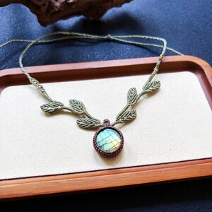 Puede incluir: Un collar de macram&eacute; con una piedra de labradorita redonda engastada en cord&oacute;n marr&oacute;n oscuro. La cadena del collar est&aacute; hecha de cord&oacute;n verde oliva claro y decorada con varios peque&ntilde;os detalles tejidos en forma de hoja a ambos lados.