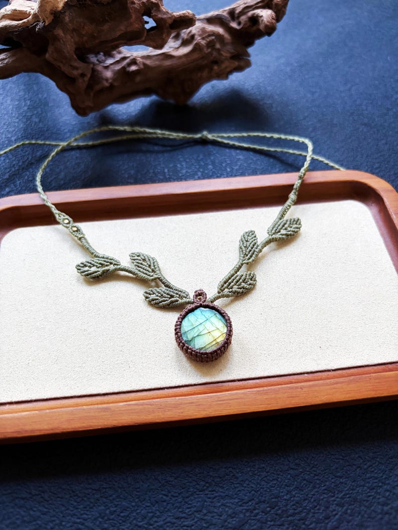 Puede incluir: Un collar de macram&eacute; con una piedra de labradorita redonda engastada en cord&oacute;n marr&oacute;n oscuro. La cadena del collar est&aacute; hecha de cord&oacute;n verde oliva claro y decorada con varios peque&ntilde;os detalles tejidos en forma de hoja a ambos lados.