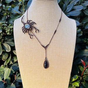 May include: A brown macrame necklace with a scorpion pendant featuring a blue-green stone and a teardrop-shaped black stone pendant. The necklace is displayed on a beige mannequin against a backdrop of green foliage.