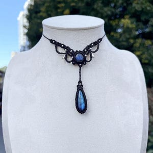 Peut inclure: Un collier en macramé noir avec un pendentif central en pierre précieuse bleue et une goutte en forme de goutte d'eau. Le collier présente des nœuds complexes et des accents argentés, présenté sur un mannequin blanc.