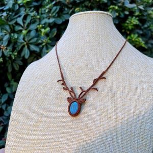 May include: A brown macrame necklace with a deer antler design. The pendant features a blue oval stone set within the macrame. The necklace is displayed on a beige mannequin head against a backdrop of green foliage.