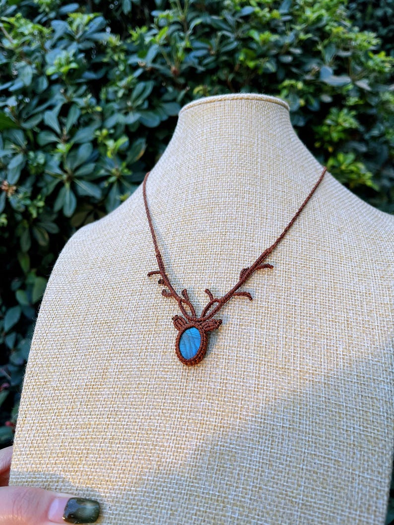 May include: A brown macrame necklace with a deer antler design. The pendant features a blue oval stone set within the macrame. The necklace is displayed on a beige mannequin head against a backdrop of green foliage.