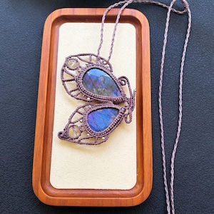 May include: A butterfly-shaped pendant necklace crafted with brown macrame cord, featuring two teardrop-shaped labradorite stones with blue and purple iridescence, accented by small clear beads and a long, braided adjustable cord with bead ends.