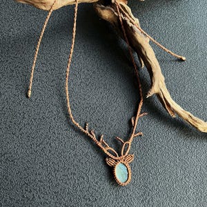 May include: A handmade macrame necklace featuring a deer antler design. The pendant has a light blue oval stone. The necklace is made of brown cord and is displayed on a dark gray surface with a piece of driftwood.