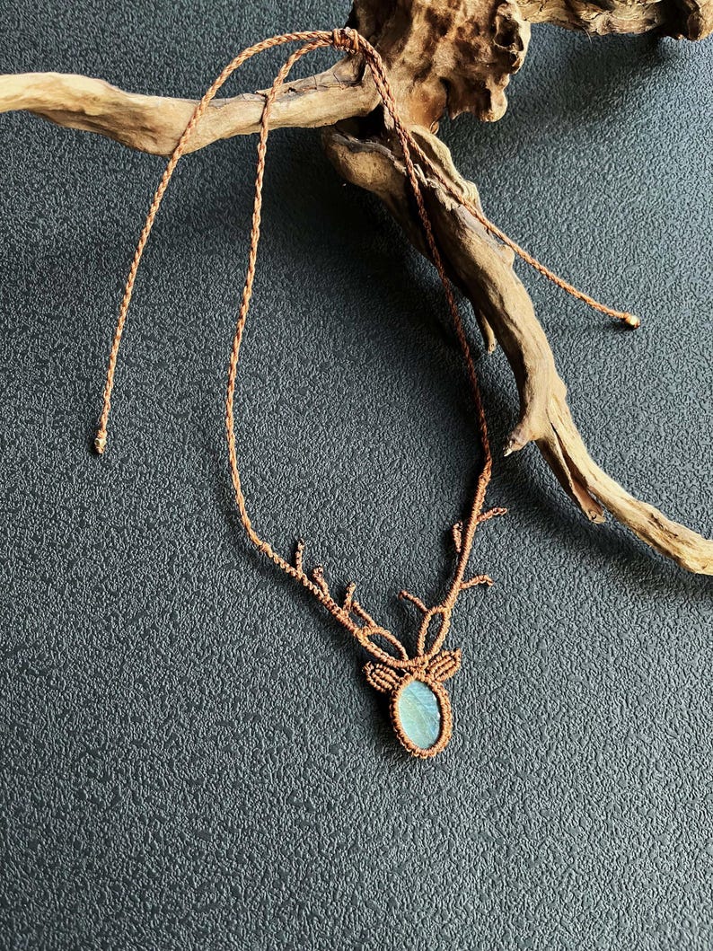 May include: A handmade macrame necklace featuring a deer antler design. The pendant has a light blue oval stone. The necklace is made of brown cord and is displayed on a dark gray surface with a piece of driftwood.
