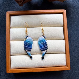May include: A pair of macrame earrings featuring a wing-shaped design with a gradient of white, light blue, and dark blue threads. Each earring is suspended from a gold-toned hook and finished with a small black bead at the bottom tip.
