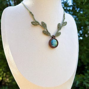 Puede incluir: Un collar de macram&eacute; con una piedra de labradorita redonda e iridiscente en tonos azules y verdes, engastada en cord&oacute;n marr&oacute;n. La cadena est&aacute; hecha de cord&oacute;n verde claro y decorada con varios peque&ntilde;os detalles tejidos en forma de hoja.