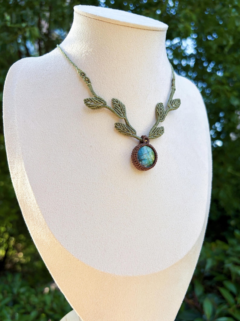 Puede incluir: Un collar de macram&eacute; con una piedra de labradorita redonda e iridiscente en tonos azules y verdes, engastada en cord&oacute;n marr&oacute;n. La cadena est&aacute; hecha de cord&oacute;n verde claro y decorada con varios peque&ntilde;os detalles tejidos en forma de hoja.
