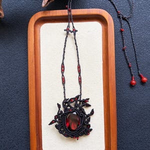 May include: A macrame necklace featuring a large, oval-shaped faceted red gemstone centrepiece surrounded by intricate black cord knotting. The cord is accented with small red beads and terminates in two long strands with larger red bead ends.