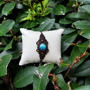 May include: A handmade macrame bracelet with a central turquoise stone and brown cord. The bracelet is displayed on a small white pillow, surrounded by green foliage. The intricate knotwork and vibrant stone create a unique accessory.