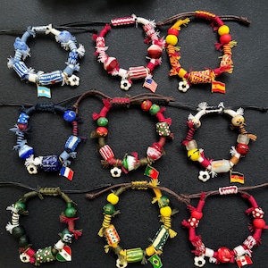 May include: A collection of nine handmade bracelets featuring colorful beads, wrapped cord, small soccer ball charms, and miniature national flag pendants. Each bracelet is constructed with adjustable dark cord and displays a unique combination of patterned beads in various shapes and sizes, accented with frayed fabric details and distinct country flags.