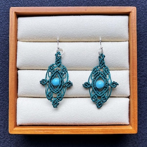 Puede incluir: Un par de pendientes de macramé turquesa con una cuenta turquesa en el centro. Los pendientes se exhiben en una caja de madera con un interior de tela blanca. Los pendientes tienen un diseño geométrico.