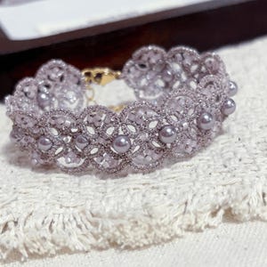 May include: A lavender-toned bracelet crafted with intricate tatting lace and small, round, lustrous beads integrated into the pattern. The bracelet features a delicate, scalloped edge design and is secured with a gold-toned metal lobster clasp.